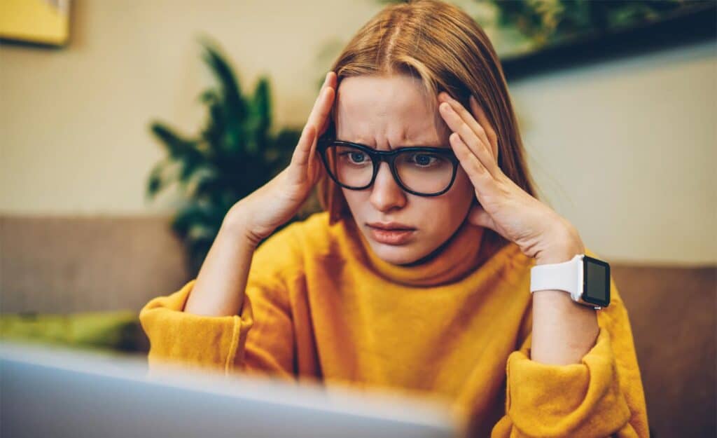 A person with glasses and a yellow sweater looks stressed while holding their head in front of a laptop.