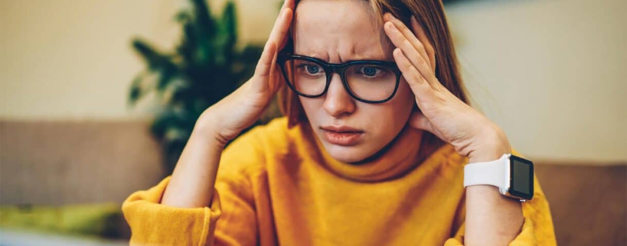 A person with glasses and a yellow sweater looks stressed while holding their head in front of a laptop.