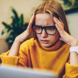 A person with glasses and a yellow sweater looks stressed while holding their head in front of a laptop.