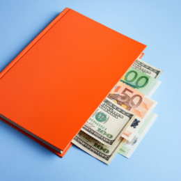 An orange book on a blue background with various paper currencies, including dollars and euros, peeking out from its pages.