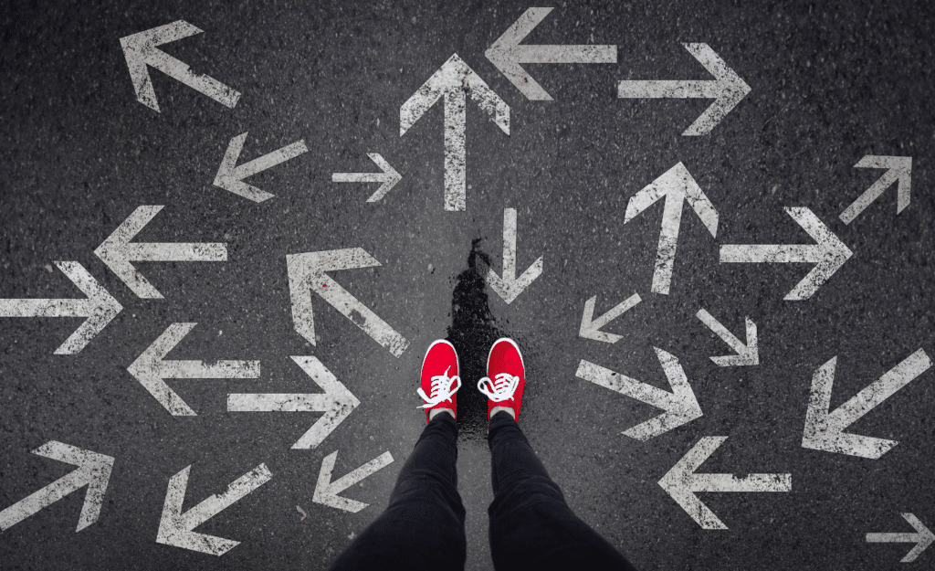 Feet with red shoes standing on pavement with multiple arrows pointing in multiple directions. This image illustrates the sometimes confusing world of AI content creation, that needs rules for AI.